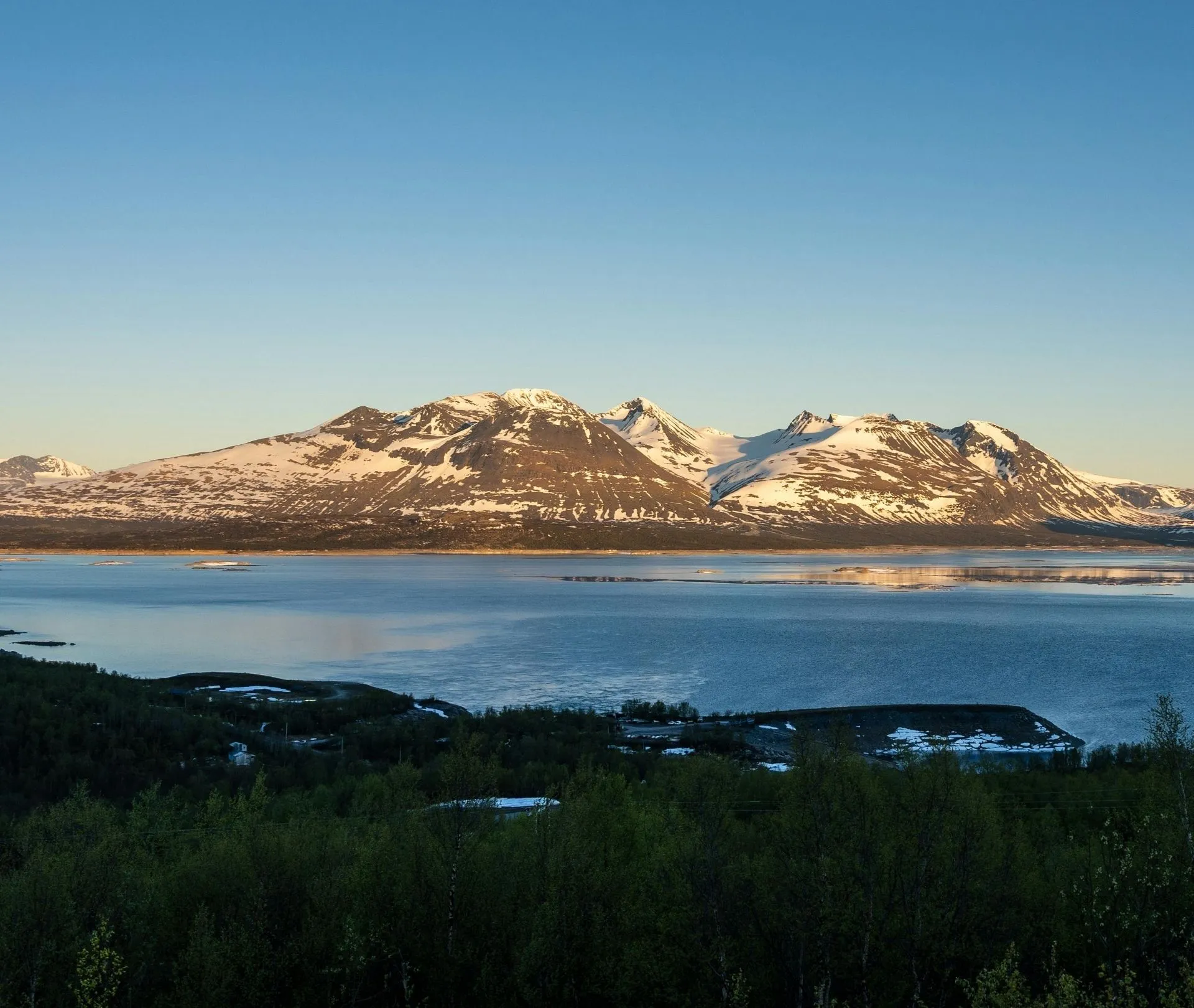 Snöklädda fjäll och klarblå sjö i Gällivare under kvällssol.